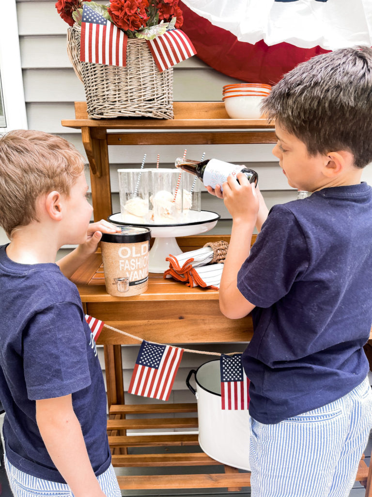Two young boys in navy t-shirts in front of 4th of July red white and blue decorated table with root beer floats one boy is pouring root bear with patriot cable