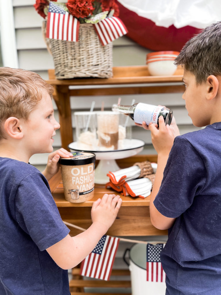 Two young boys in navy t-shirts in front of 4th of July red white and blue decorated table with root beer floats one boy is pouring root bear with patriot cable