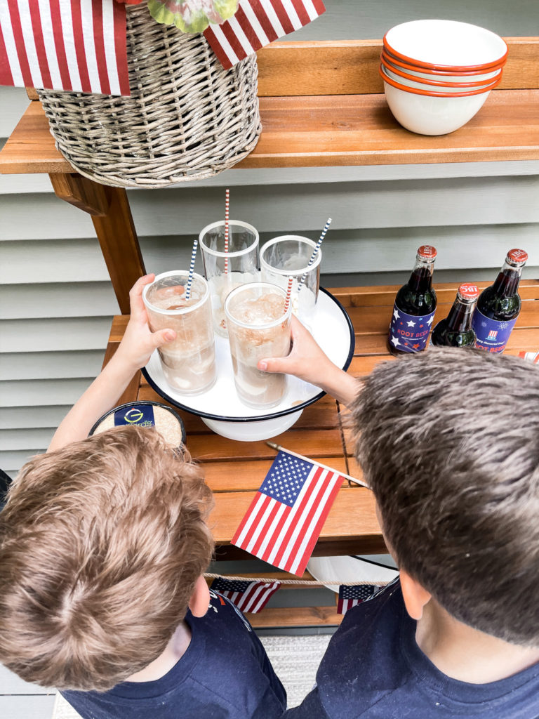 Two young boys in navy t-shirts in front of 4th of July red white and blue decorated table with root beer floats in hand