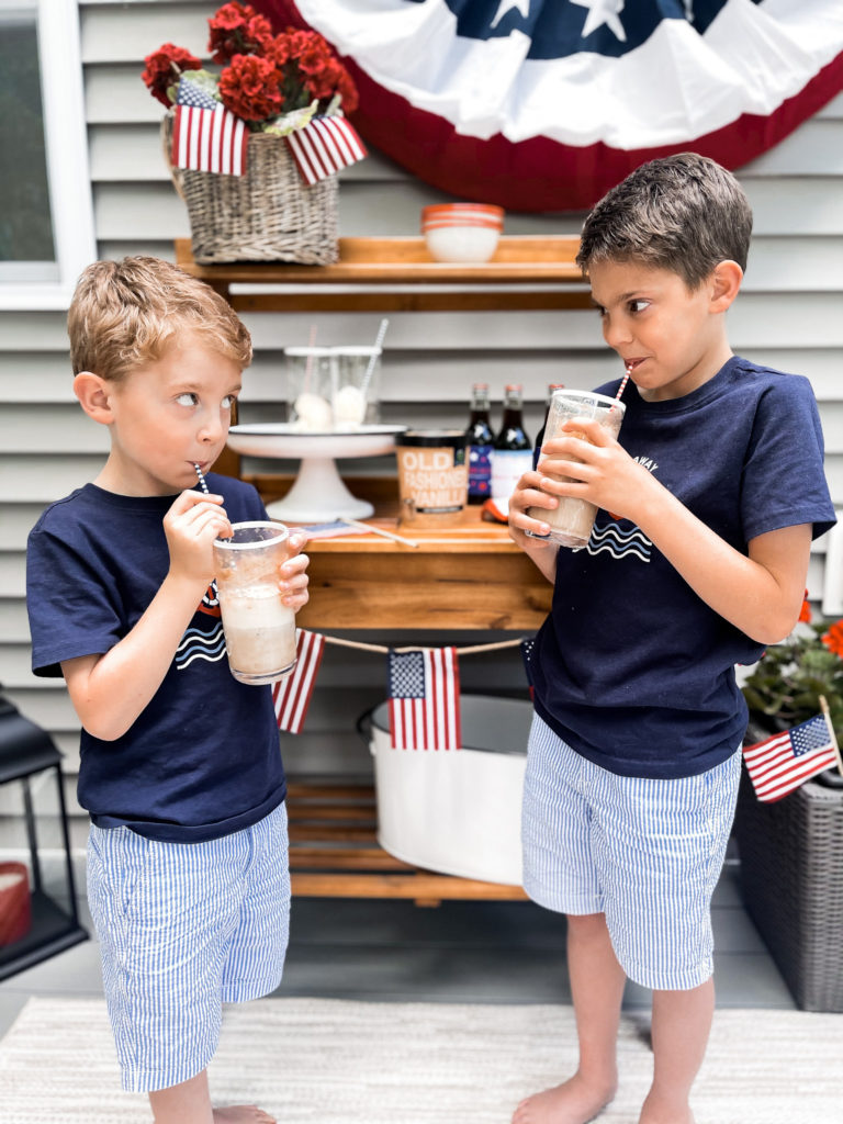 Two young boys in navy t-shirts in front of 4th of July red white and blue decorated table with root beer floats in hand