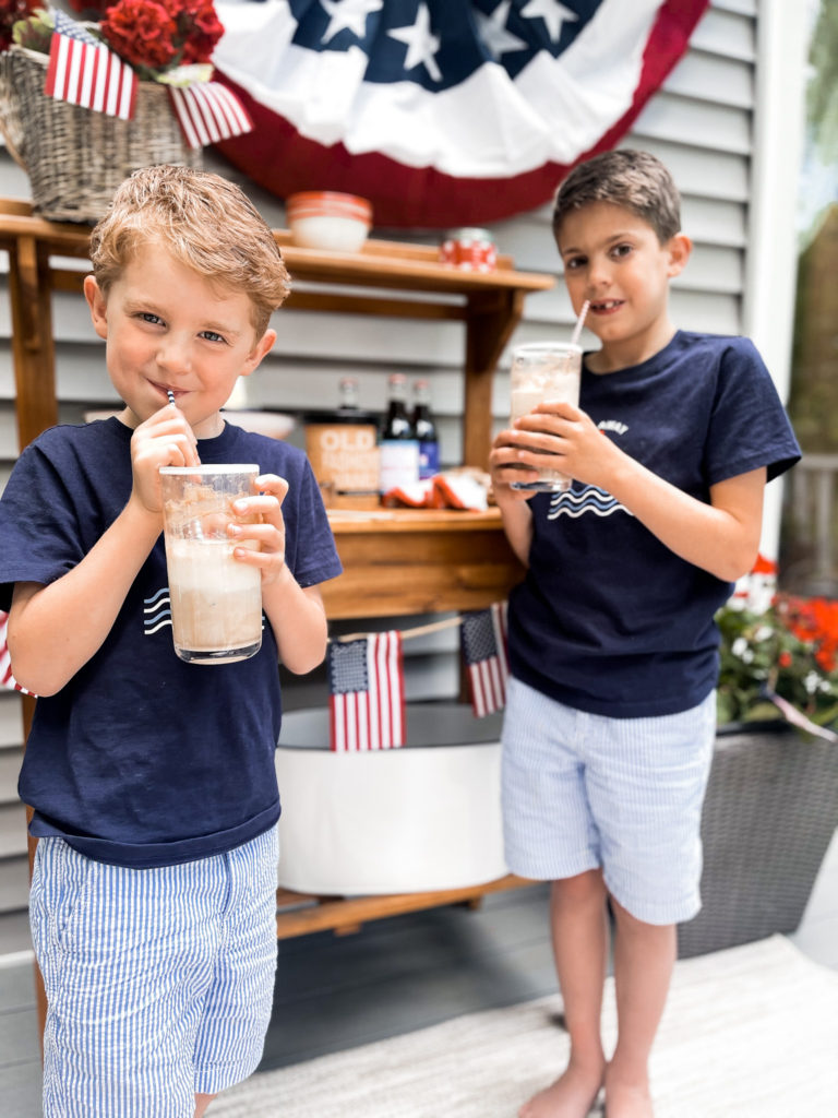 Two young boys in navy t-shirts in front of 4th of July red white and blue decorated table with root beer floats in hand