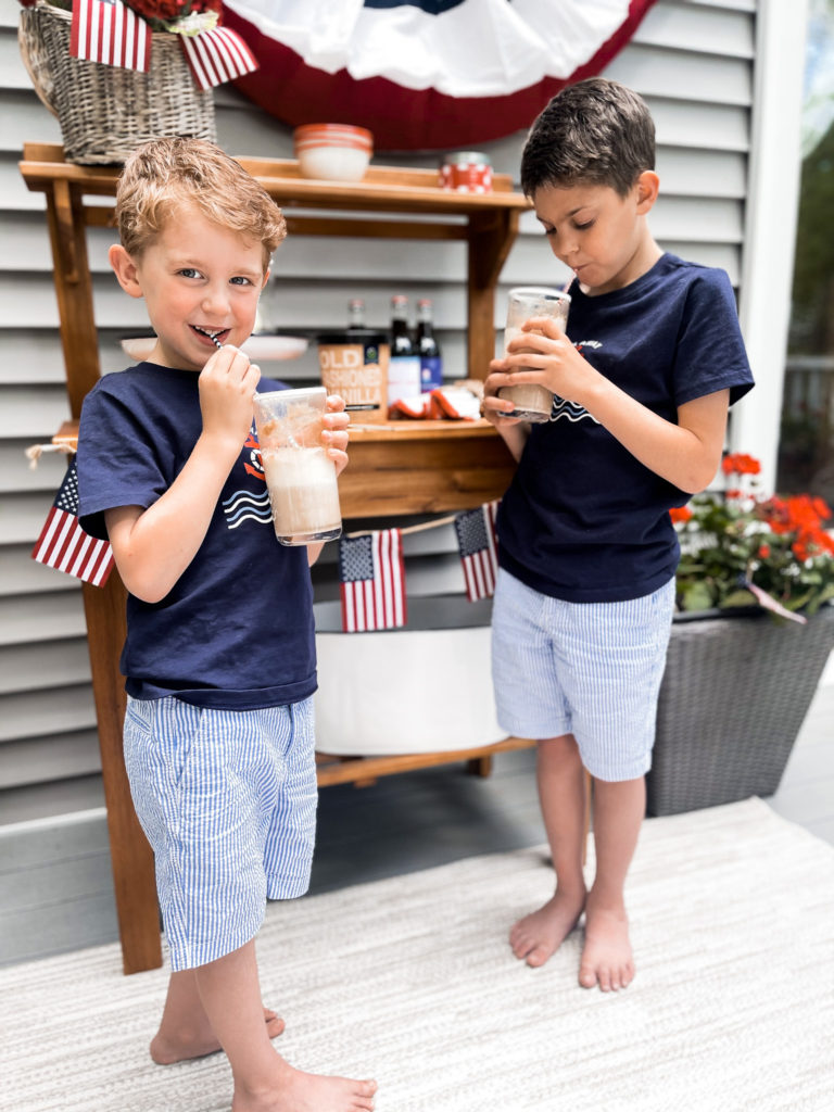 Two young boys in navy t-shirts in front of 4th of July red white and blue decorated table with root beer floats in hand