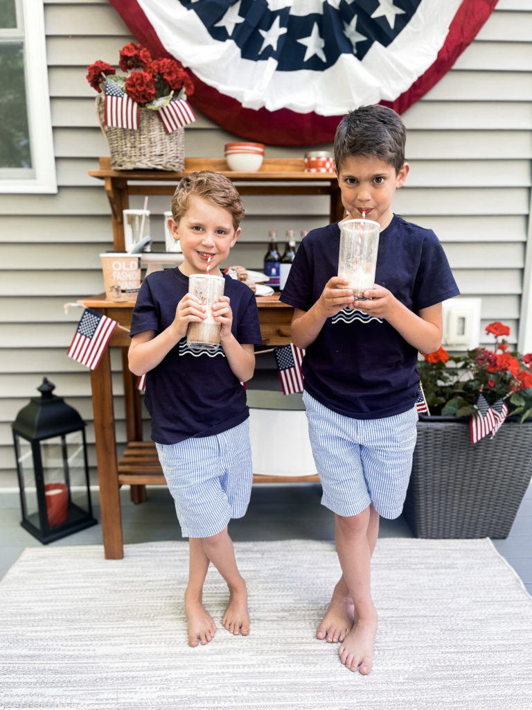 Two young boys in navy t-shirts in front of 4th of July red white and blue decorated table with root beer floats in hand